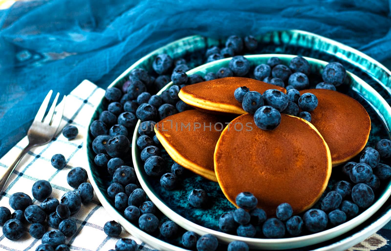 selective focus. pancakes with blueberries on a blue plate. Lots of fresh blueberries scattered around. Delicious dessert with berries for breakfast. High quality photo