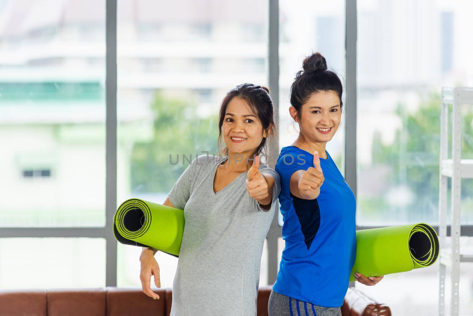 Asian adult and young woman smiling holding a yoga mat after yoga and exercise and show thumb up sign. Portrait of happy beautiful female hold yoga mat indoor studio, sport healthy workout concept