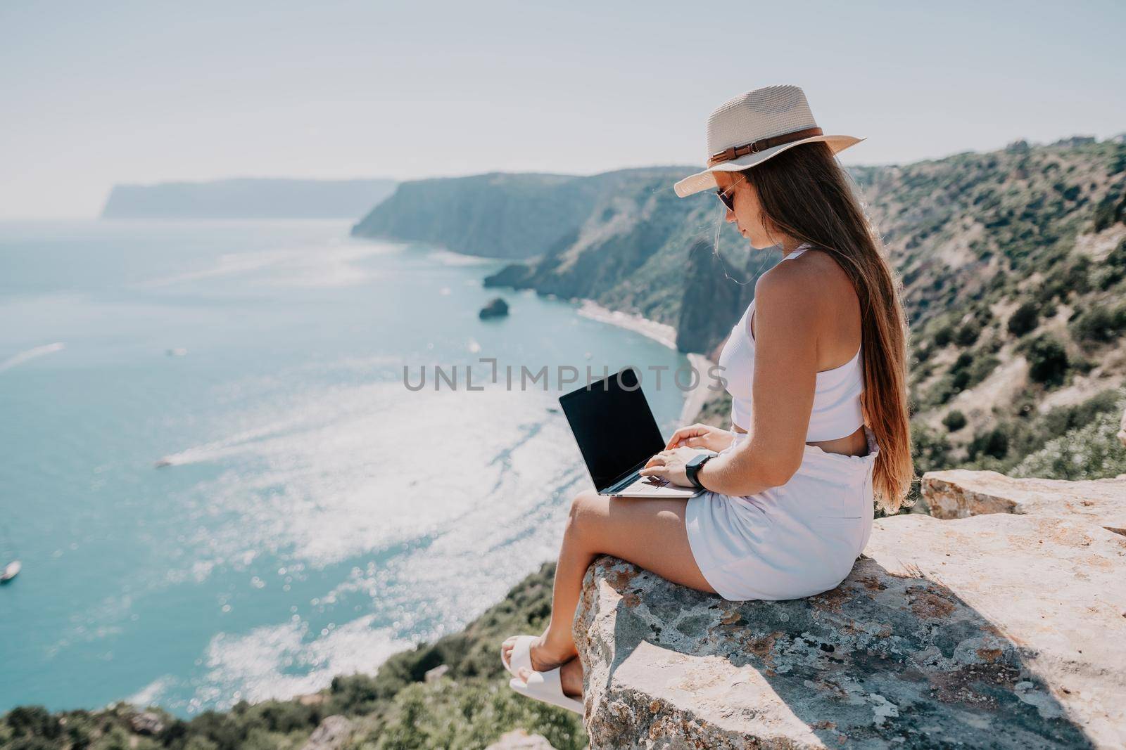 Successful business woman in yellow hat working on laptop by the sea. Pretty lady typing on computer at summer day outdoors. Freelance, travel and holidays concept.