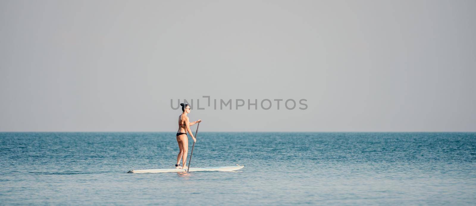 Silhouette of woman standing, surfing on SUP board, confident paddling through water surface. Idyllic sunset or sunrise. Sports active lifestyle at sea or river.