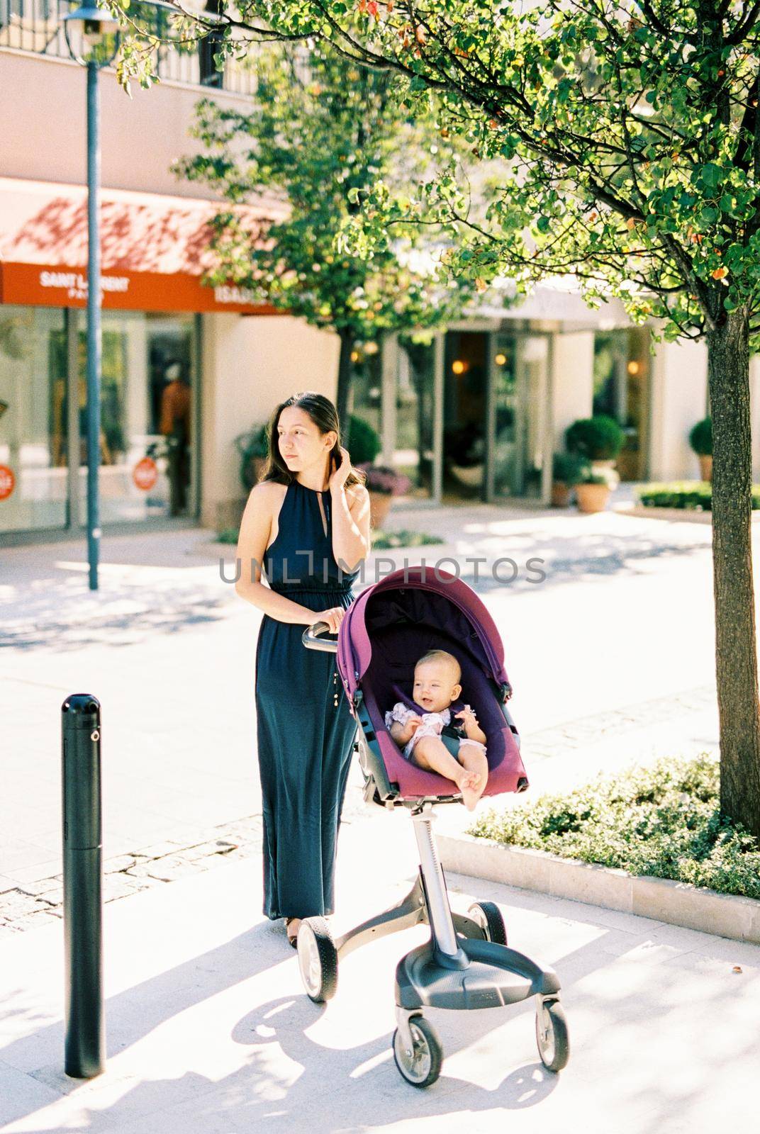 Mom with a baby in a stroller stands on the sidewalk and looks away. High quality photo