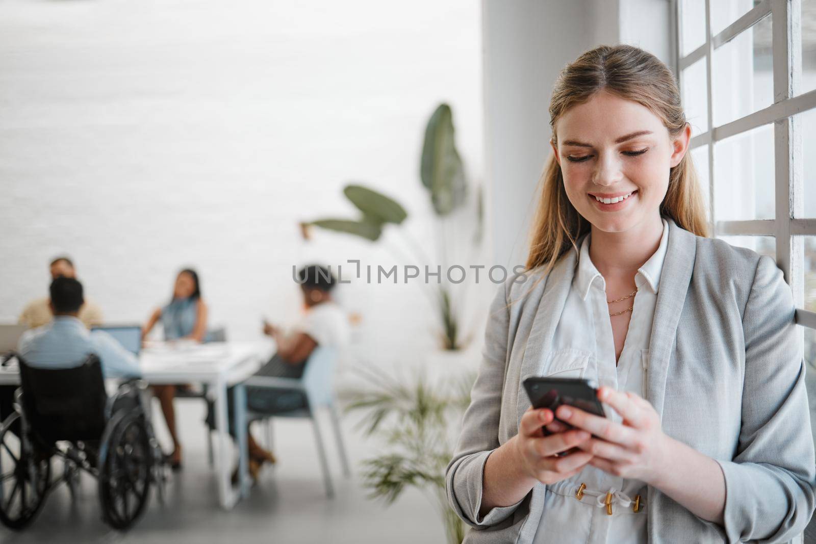 Phone, networking and communication with a business woman on social media in an office for a meeting with a team in the background. Advertising, marketing and teamwork with a worker typing a message by YuriArcurs