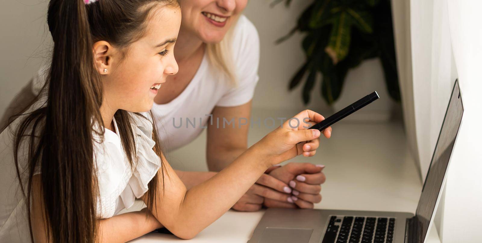 Homeschool little young girl learning online class from school teacher by digital remote internet meeting due to coronavirus pandemic. Kid looking computer and writing note with help from mother.