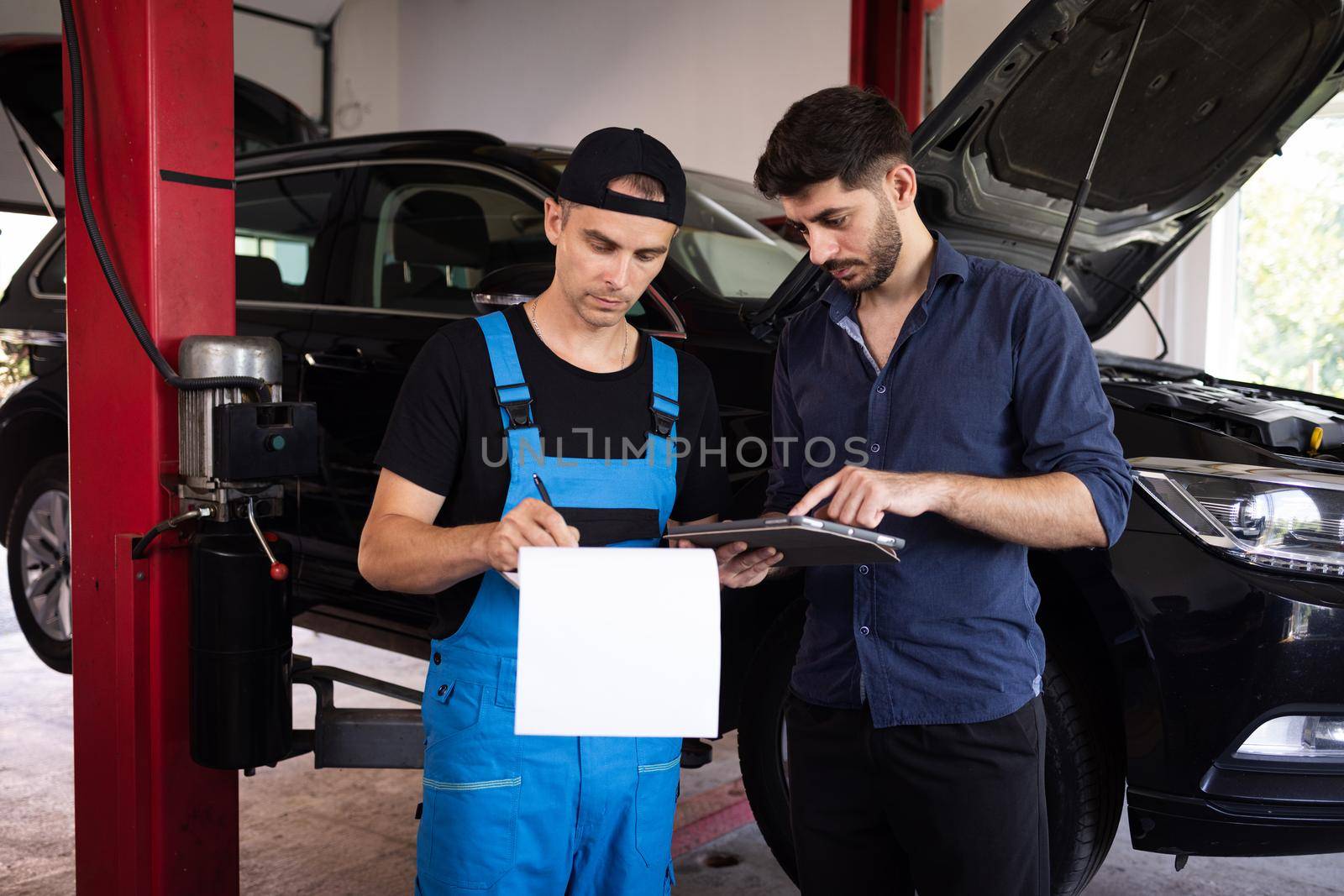Car Service Employees Inspect the Bottom and Skid Plates of the Car. Manager Checks Data on a Tablet Computer and Explains the Breakdown to Mechanic. Specialist is Showing Info on Tablet. by uflypro