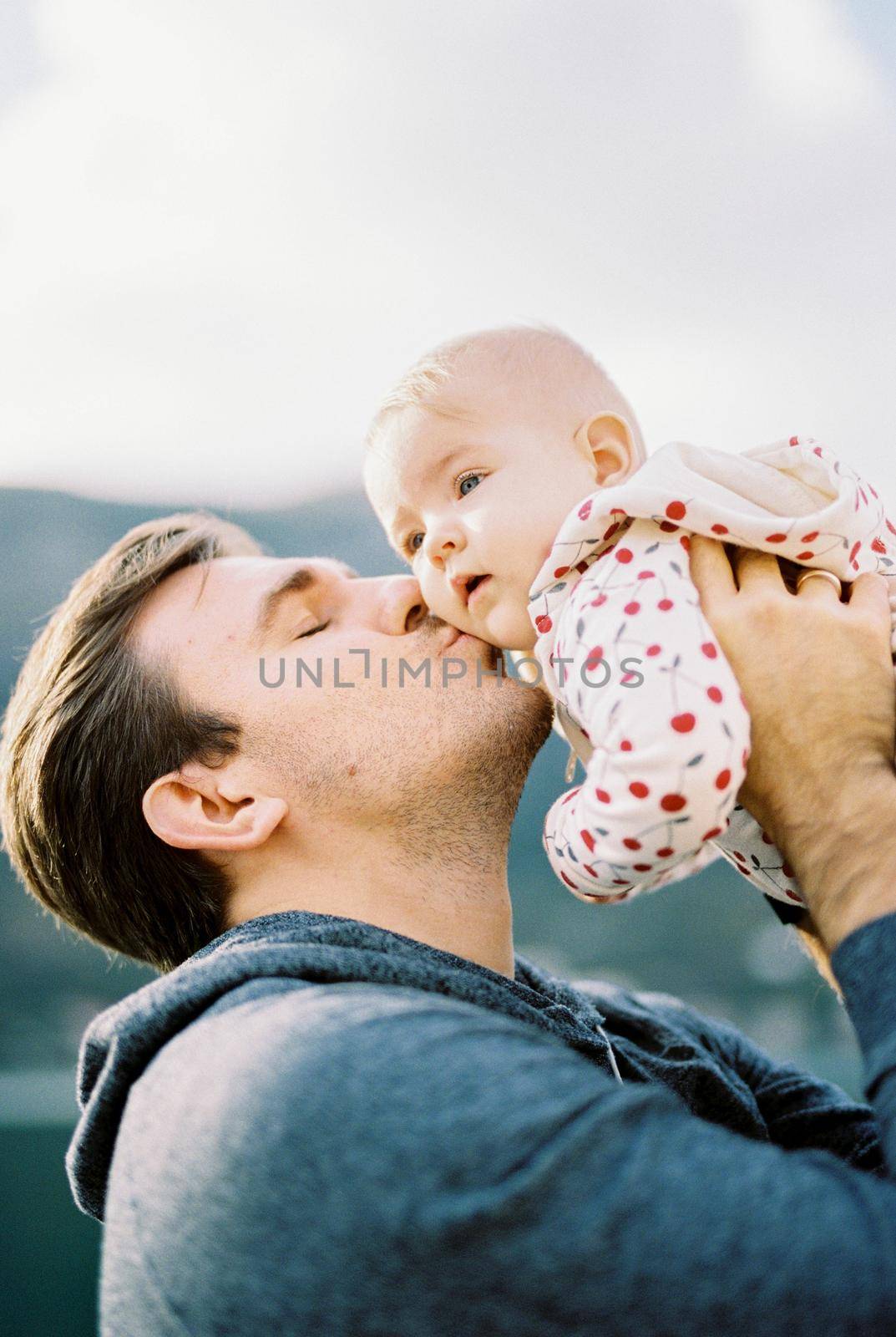 Dad lifts the baby above his head and kisses him on the cheek. Portrait by Nadtochiy