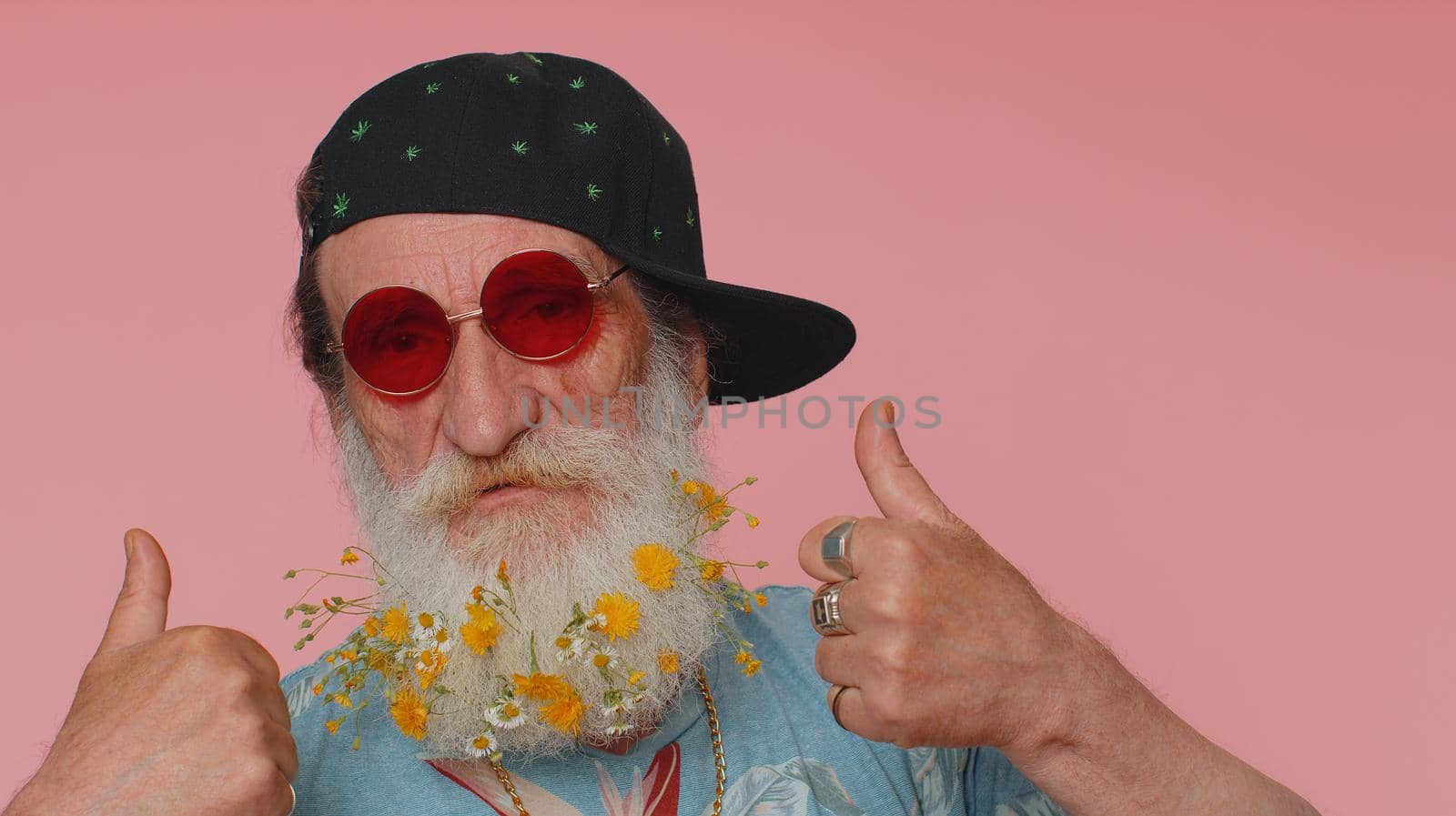 Close-up of cheerful stylish elderly man wearing sunglasses, charming smile. People emotions concept. Senior lovely mature grandfather with flowers in gray-haired beard posing isolated alone on pink