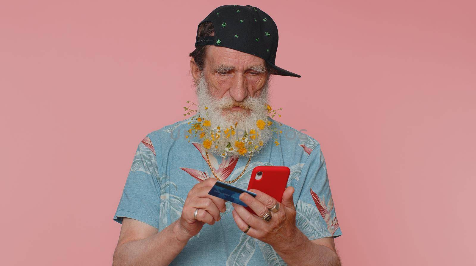 Sincere mature man customer using credit bank card and smartphone while transferring money, purchases online shopping, payment. Finance and internet. Senior grandfather alone on pink wall background