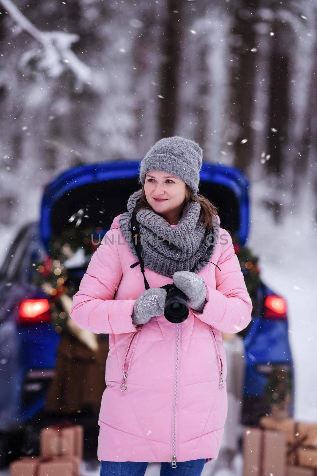 A woman in a winter snow-covered forest in the trunk of a car decorated with Christmas decor. A female photographer holds a camera in her hands.