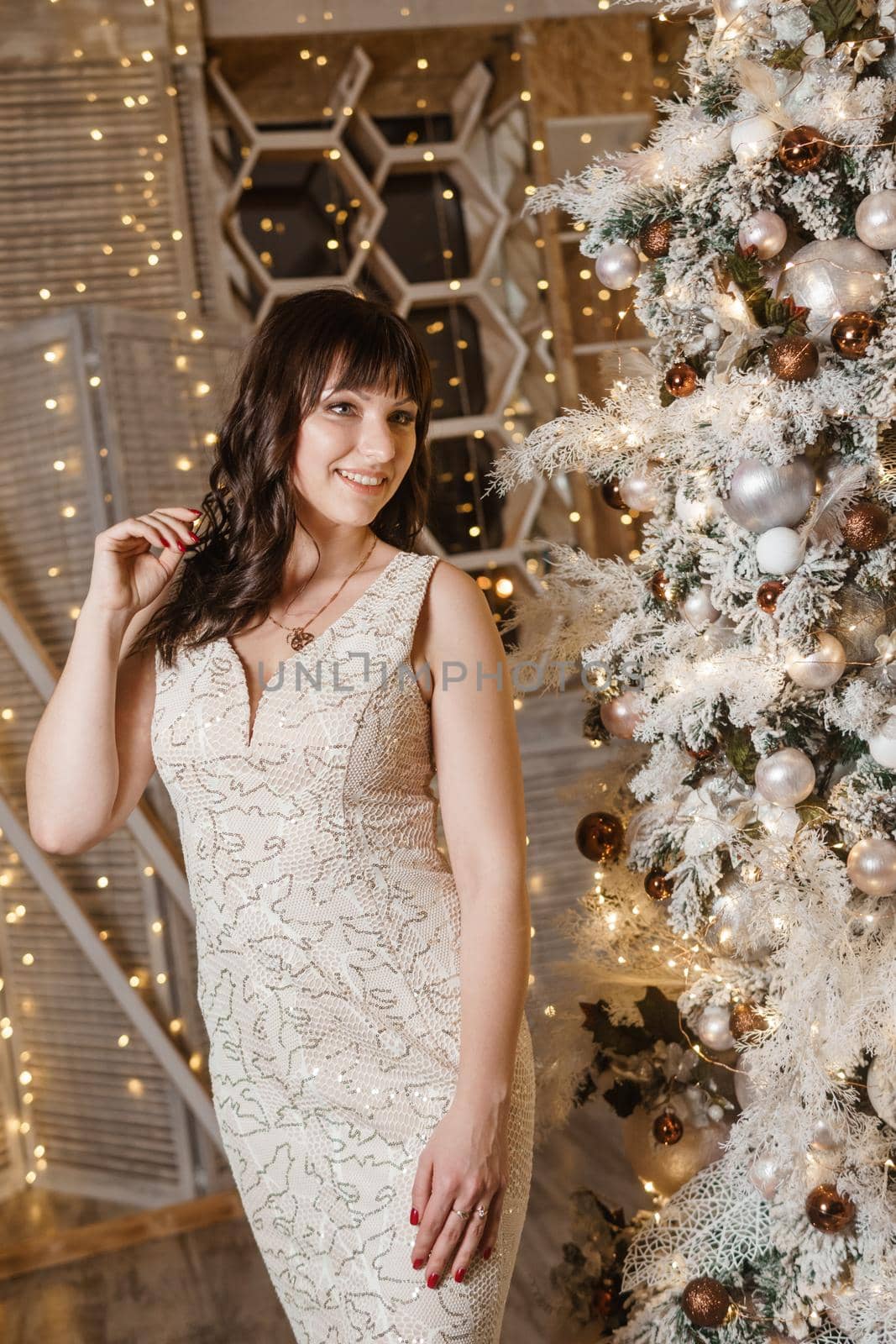 A brunette woman in a beautiful light tight dress next to a Christmas tree. The magical atmosphere of Christmas Eve on December 25. Decoration for the holiday in a light style with lots of lights and light bulbs.