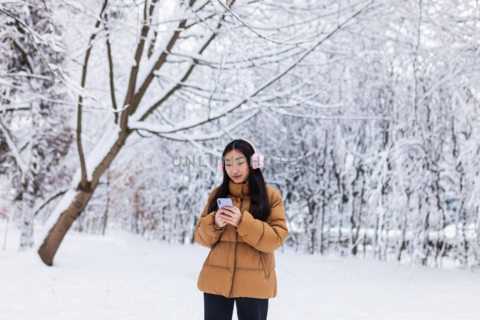 Beautiful Japanese teenager walks in the winter park uses the phone and listens to music with cute pink headphones