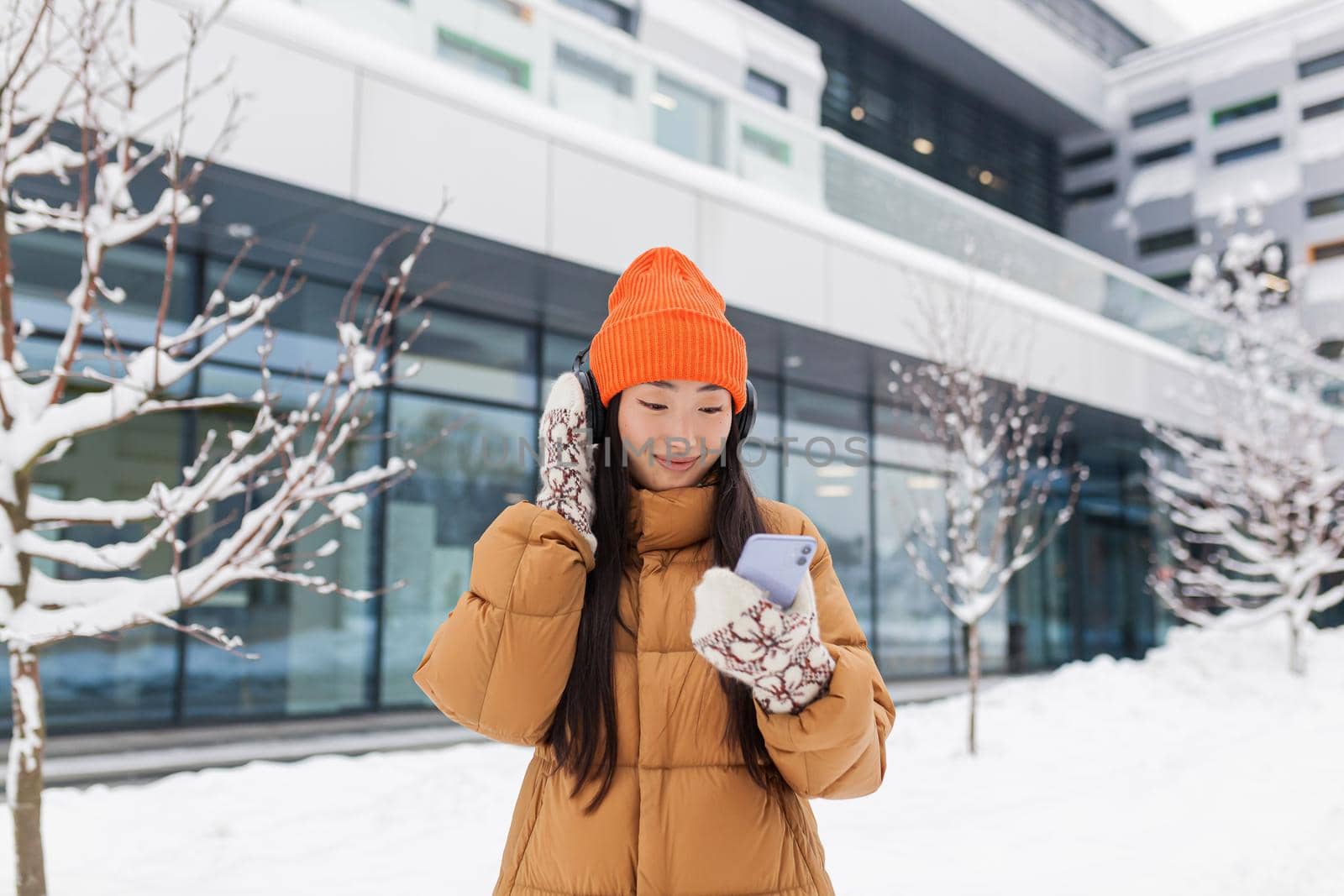 Young beautiful Asian woman listens to music from headphones while walking on a winter snowy day, uses an online application for the phone