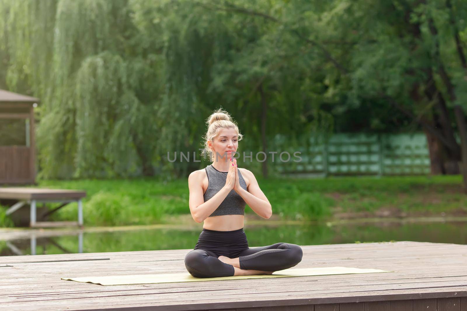 A beautiful woman in a gray top and leggings, sitting on a wooden platform by a pond in the park in summer, does yoga with her palms together. Copy space