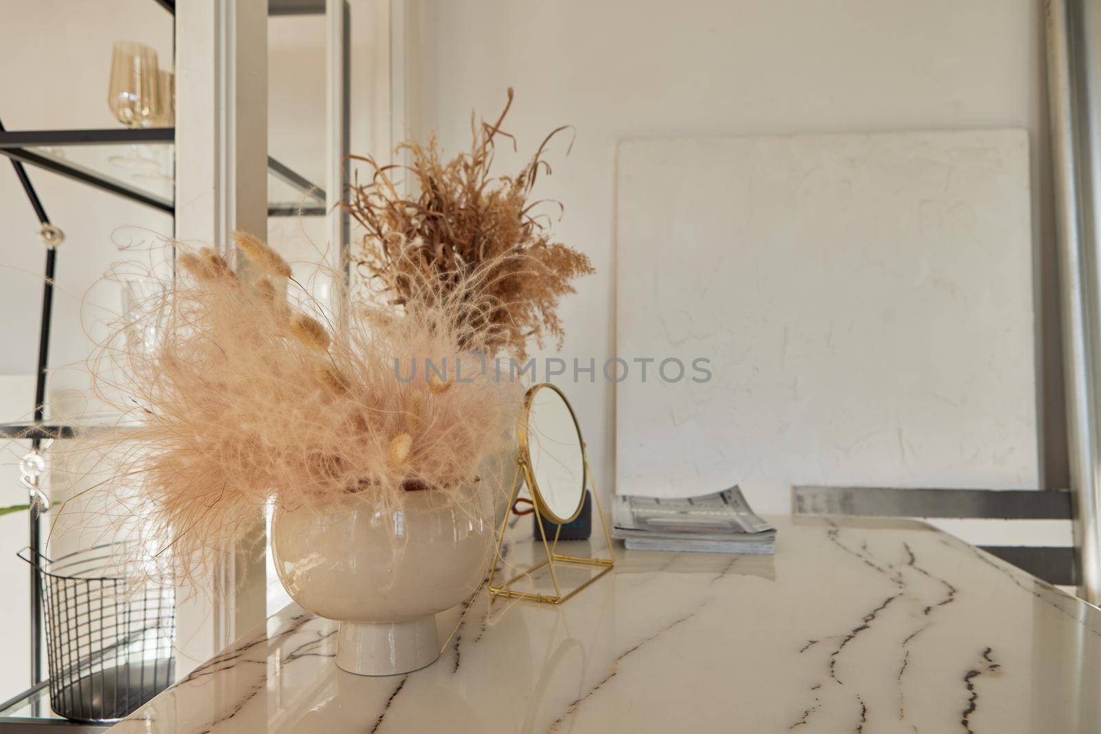 Dry decorative plants in elegant vases placed on marble table near round shaped cosmetic mirror and magazines in classic style room