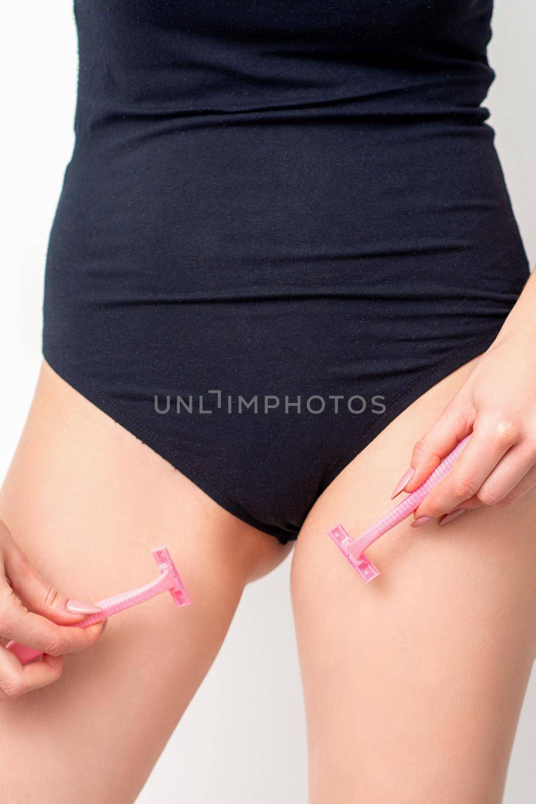 Young woman shaving crotch in bikini zone with two razors standing on white background, concept skincare and hair removal