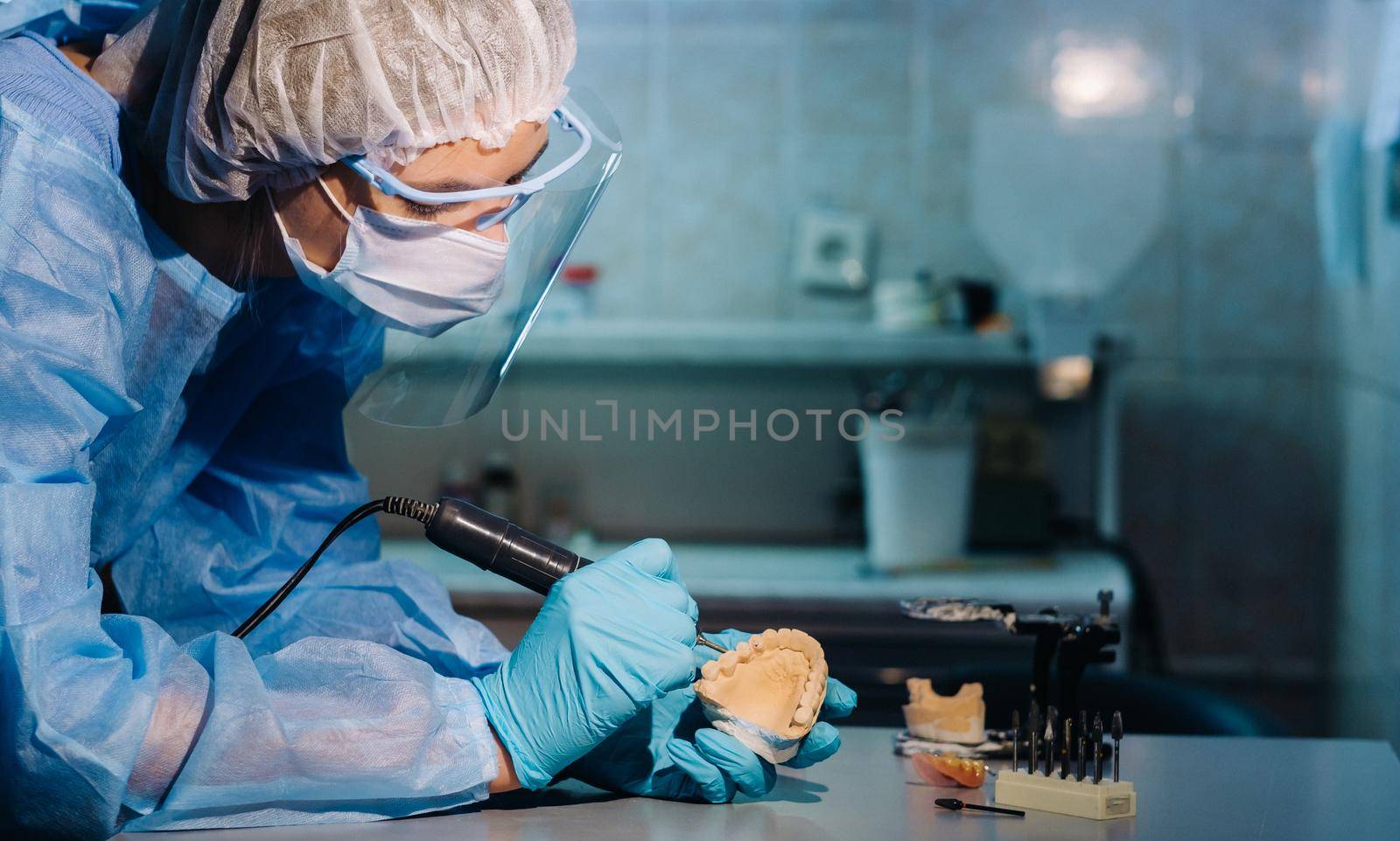 A masked and gloved dental technician works on a prosthetic tooth in his lab by Lobachad