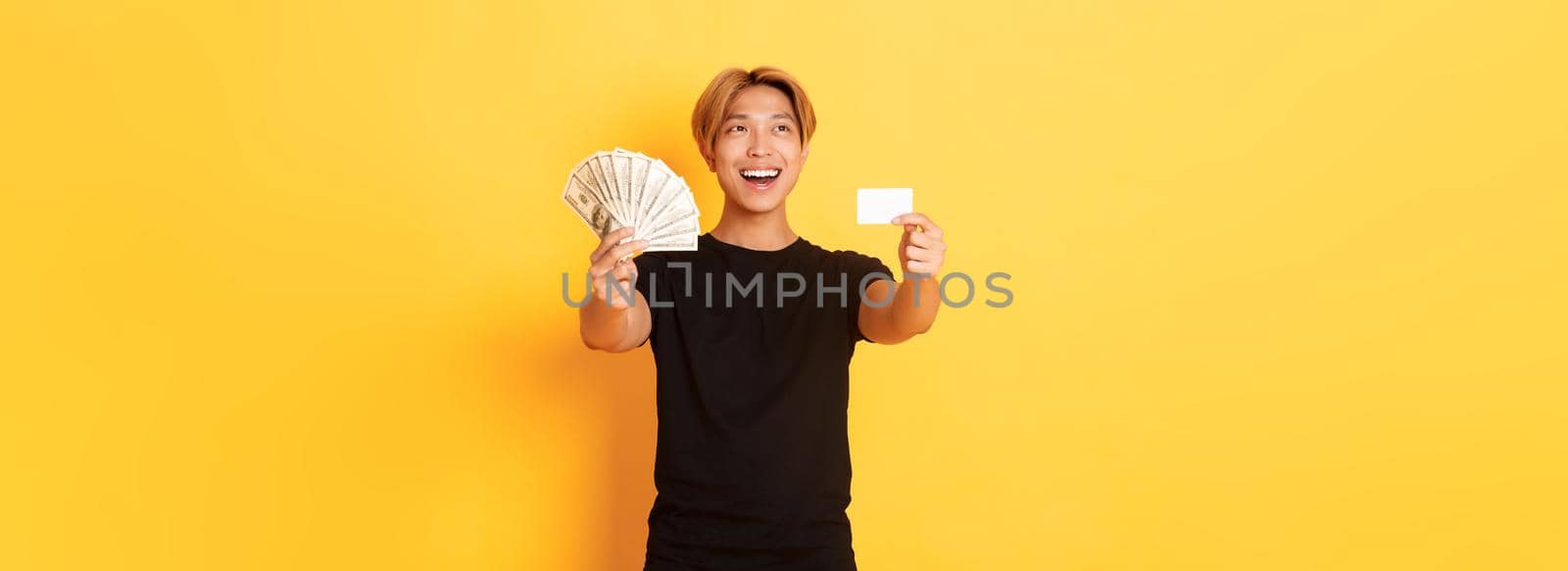 Happy good-looking asian guy looking thoughtful and pleased upper left corner while showing money and credit card, yellow background.