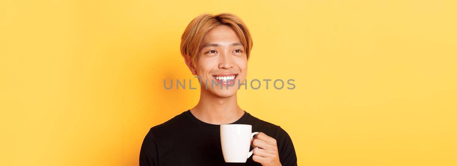 Close-up of happy handsome young asian guy with blond hair, looking dreamy and smiling while drinking coffee or tea, standing over yellow background by Benzoix