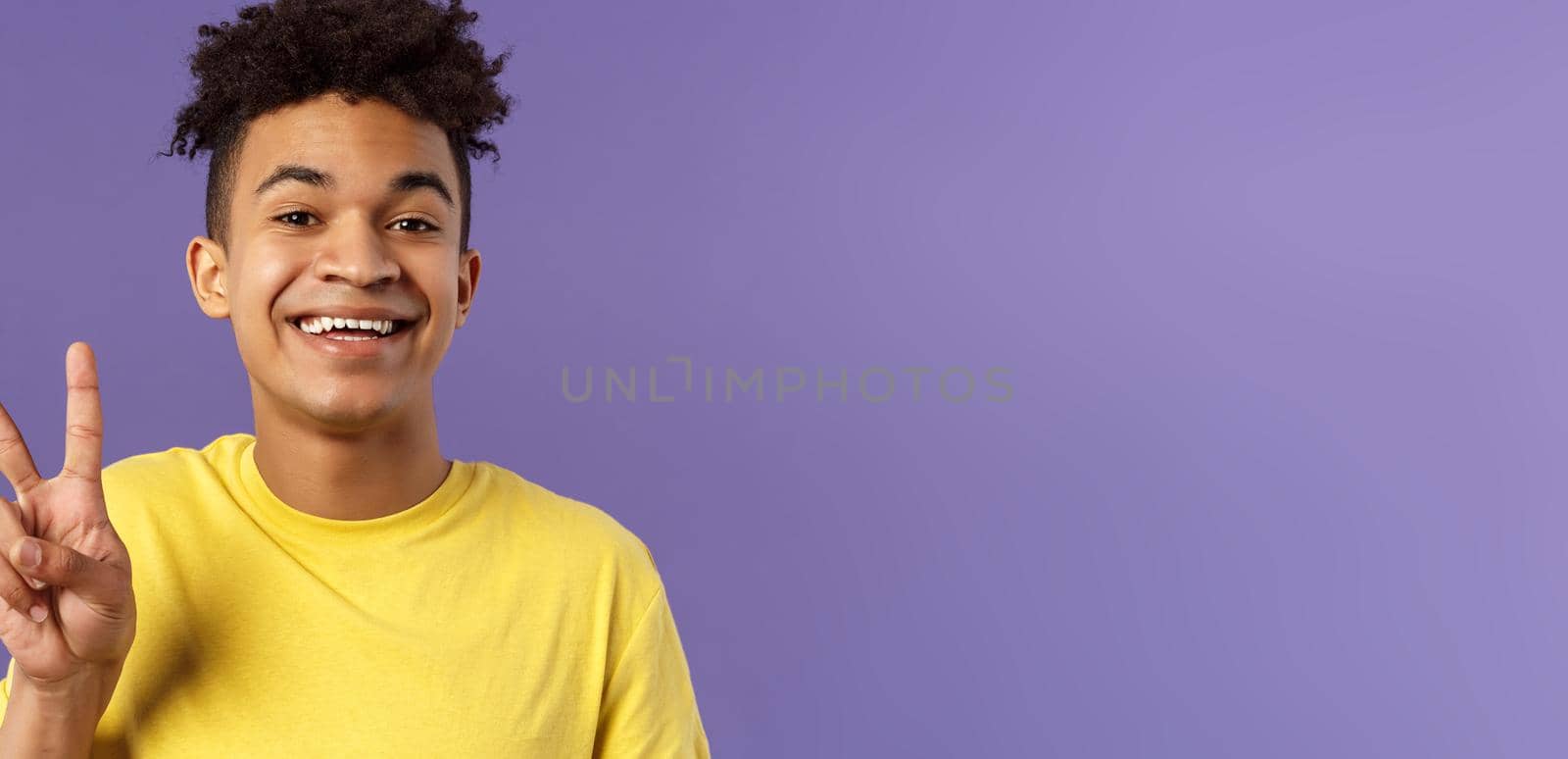 Close-up portrait of handsome upbeat young teenage guy with afro hairstyle, show peace sign and smiling, wear yellow t-shirt, staying optimistic and positive, purple background.