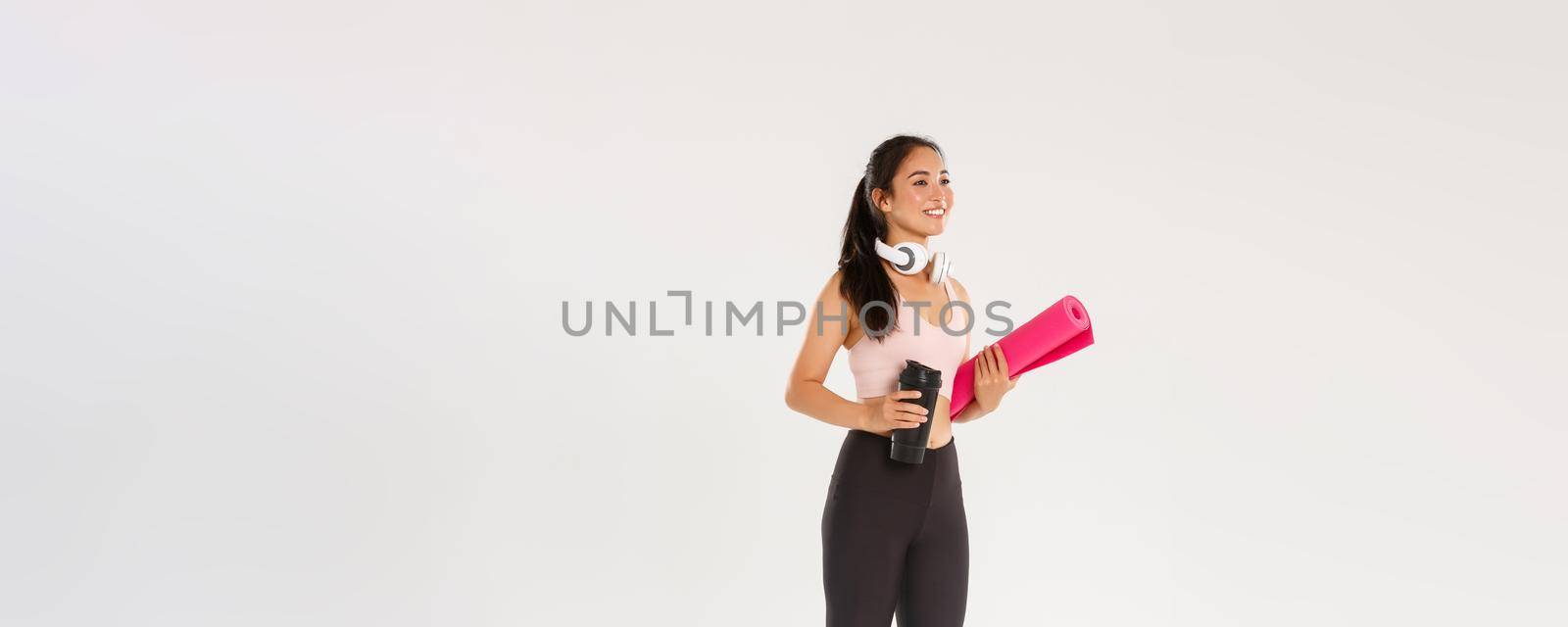 Full length side view of beautiful asian girl going for yoga classes, female athlete with headphones and water bottle smiling as carry fitness exercises rubber mat for her training in gym.