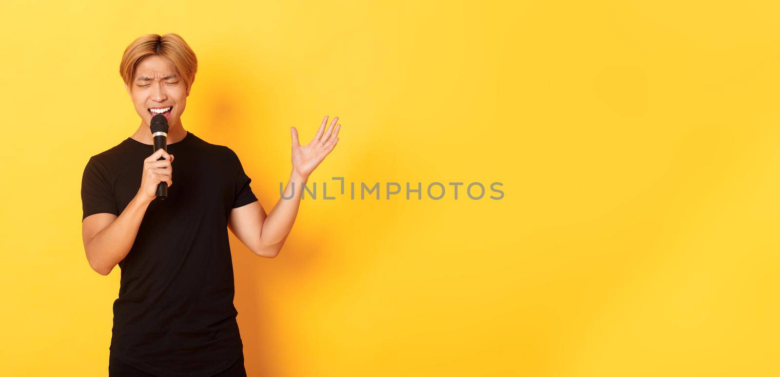 Handsome asian male singer, korean guy singing song at karaoke in microphone with passion, standing over yellow background.