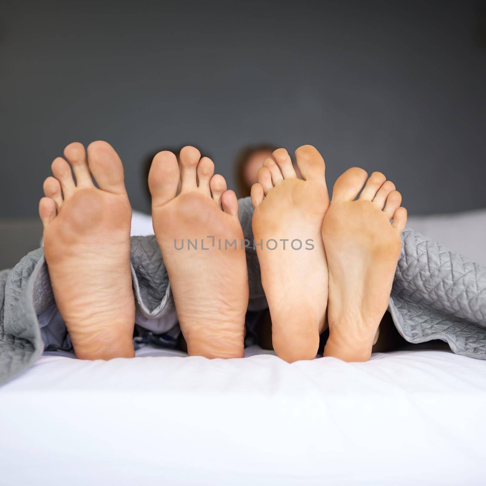 Its Sunday, put your laziest foot forward. a couples feet poking out from under the bed sheets. by YuriArcurs