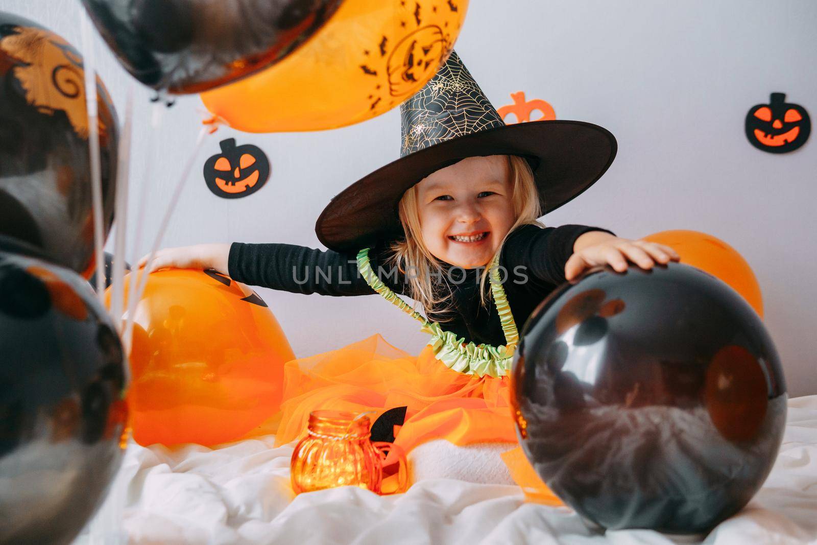 Children's Halloween - a girl in a witch hat and a carnival costume with airy orange and black balloons at home. Ready to celebrate Halloween.