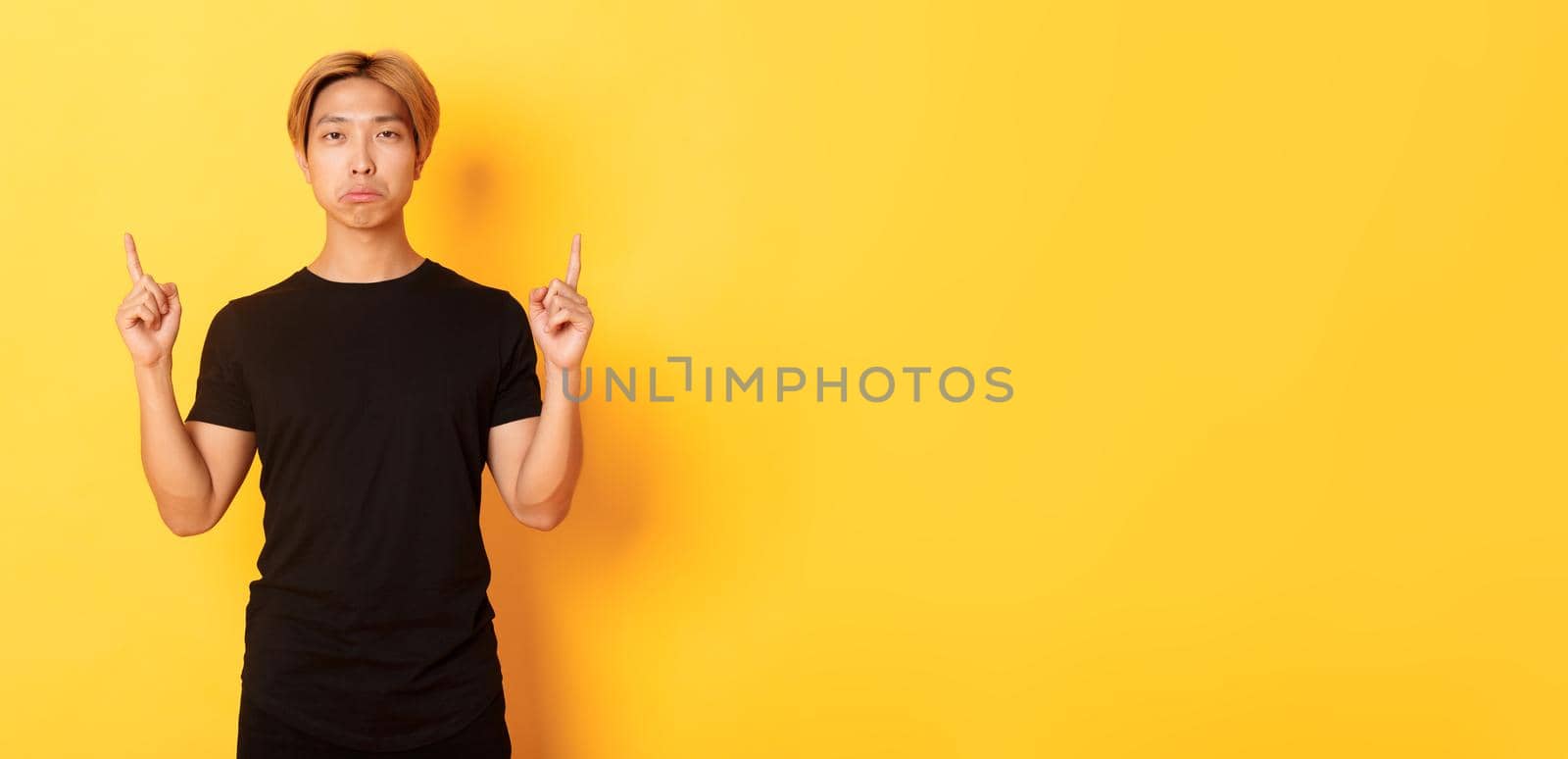 Portrait of sulking gloomy asian guy looking disappointed, pointing fingers up, yellow background.
