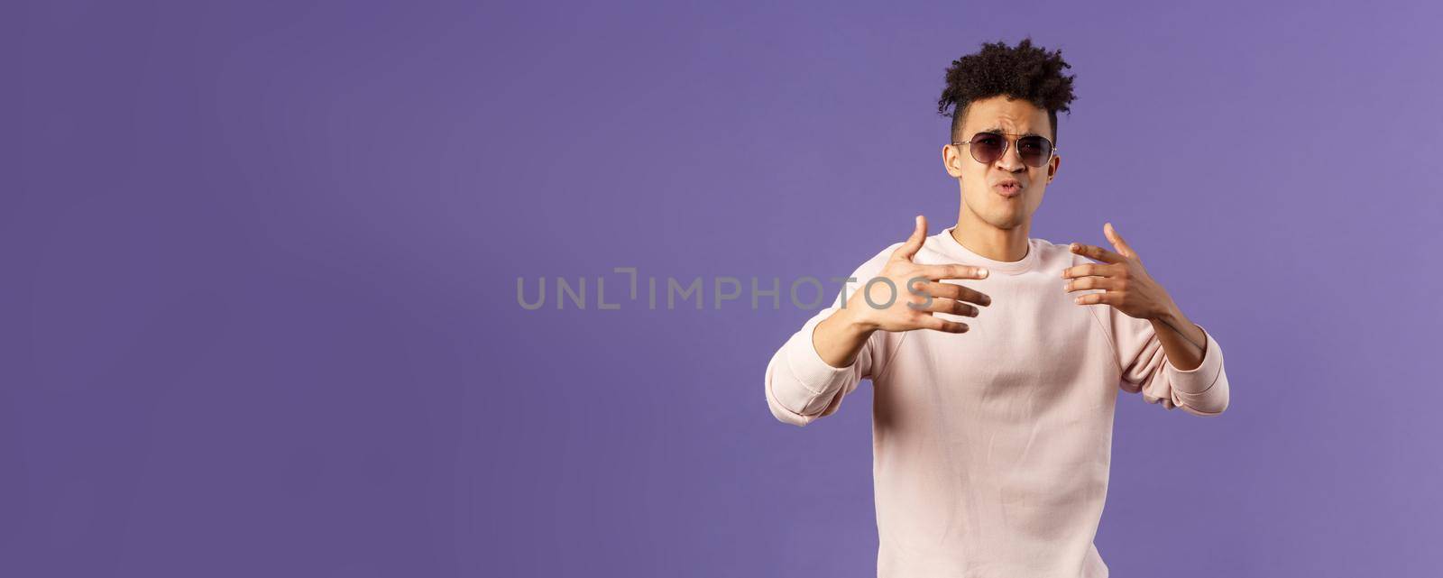 Portrait of sassy and carefree young hipster guy with dreads, wearing sunglasses dancing and gesturing while singing during rap battle, dance hip-hop on party, standing purple background.