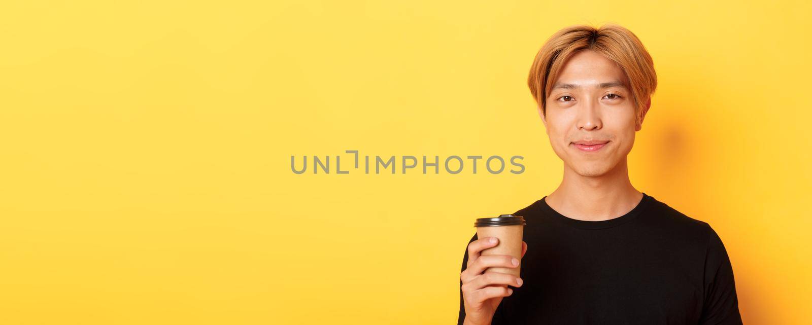 Close-up of handsome blond asian man drinking coffee and smiling pleased, standing over yellow background.
