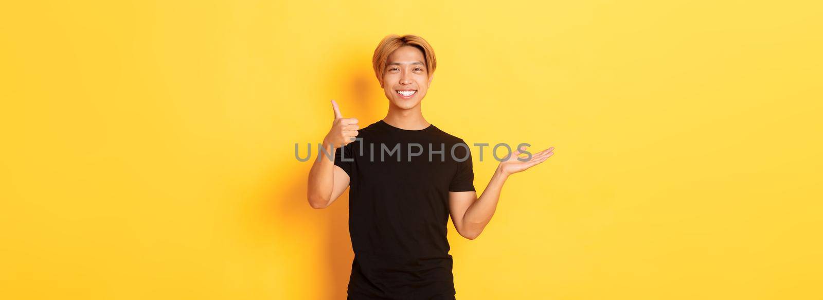 Satisfied and happy attractive korean guy smiling, showing thumbs-up in approval with rejoice, holding something on hand, standing yellow background.