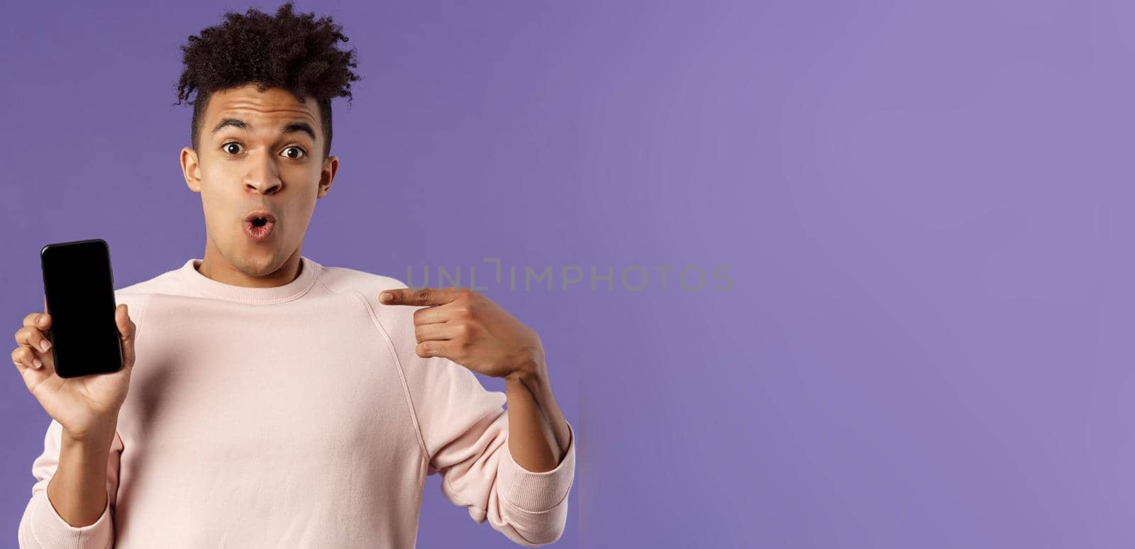 Close-up portrait of excited, cheerful young hispanic male geek talking about his device, new gadget or application, holding mobile phone, pointing at smartphone with amused face.