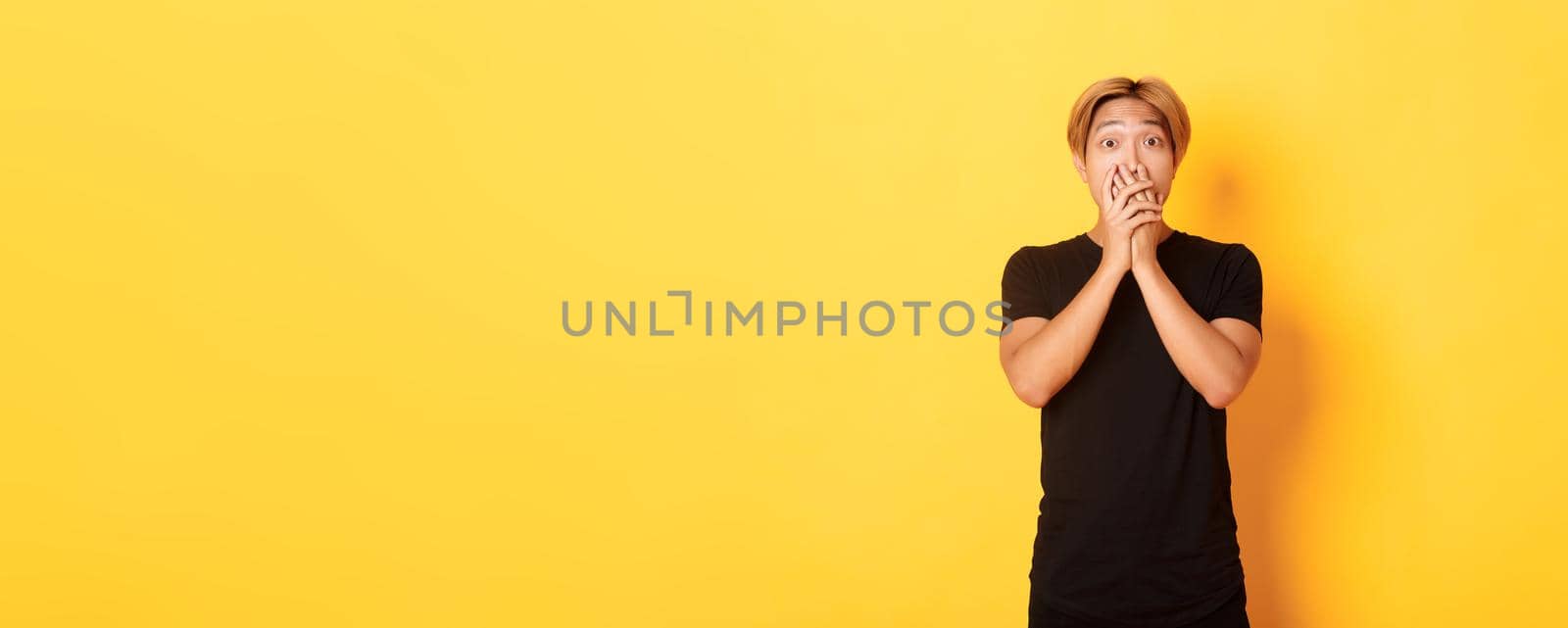 Portrait of surprised asian handsome guy with blond hair, cover mouth with hands and stare at camera impressed, yellow background.