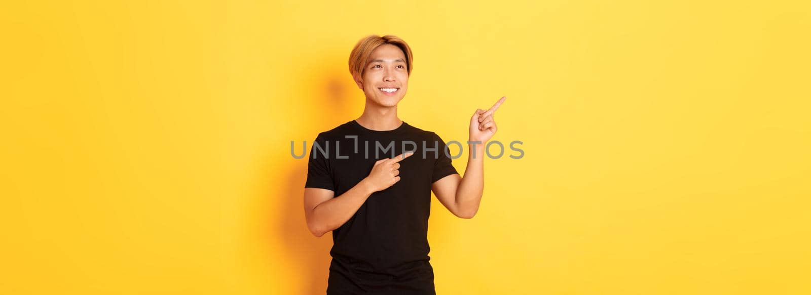 Portrait of handsome korean blond guy, pointing fingers upper left corner and looking satisfied with pleased smile, yellow background.