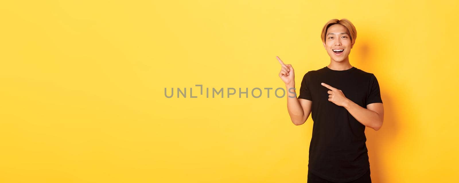 Portrait of cheerful asian man pointing fingers upper left corner, smiling and showing banner satisfied, yellow background by Benzoix