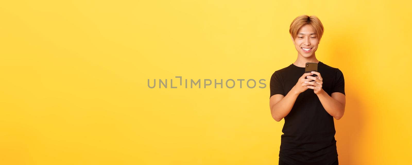 Portrait of handsome stylish asian guy with blond hair, using mobile phone and smiling, yellow background by Benzoix