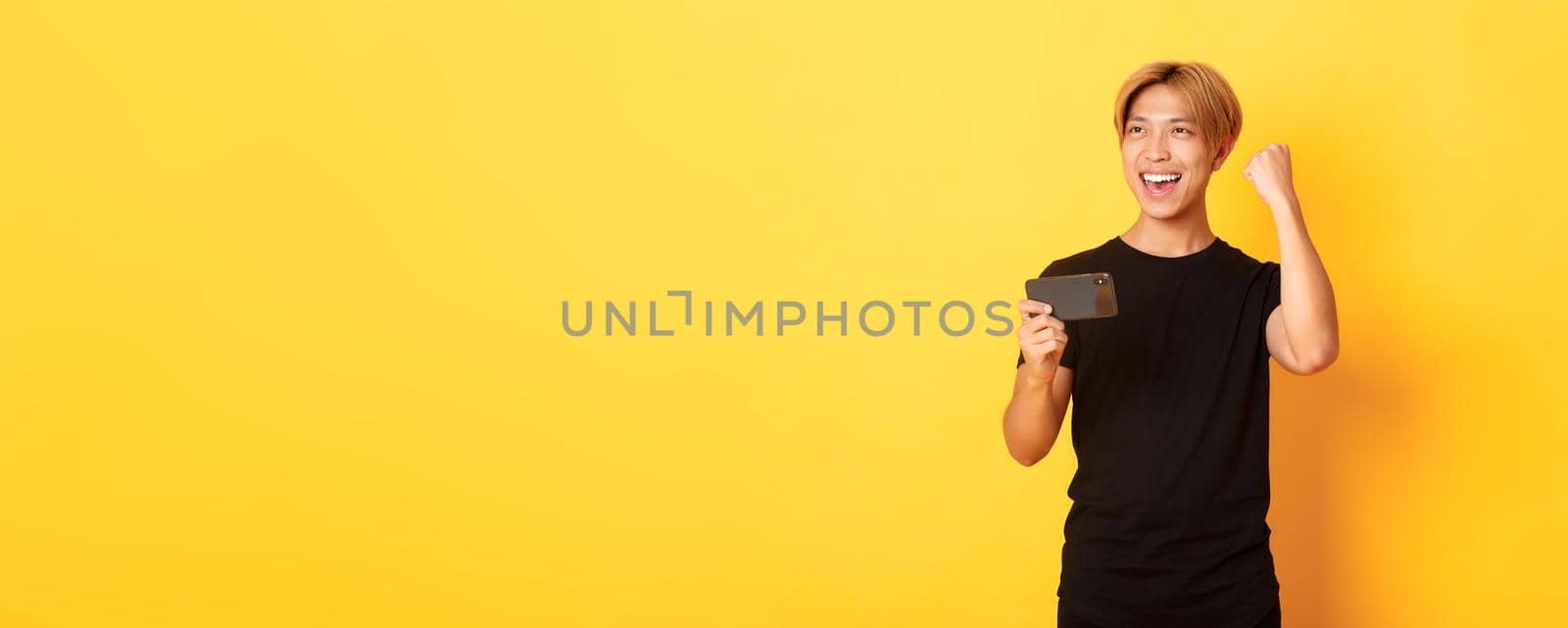 Portrait of stylish handsome asian guy playing smartphone game, rejoicing from winning, standing yellow background.