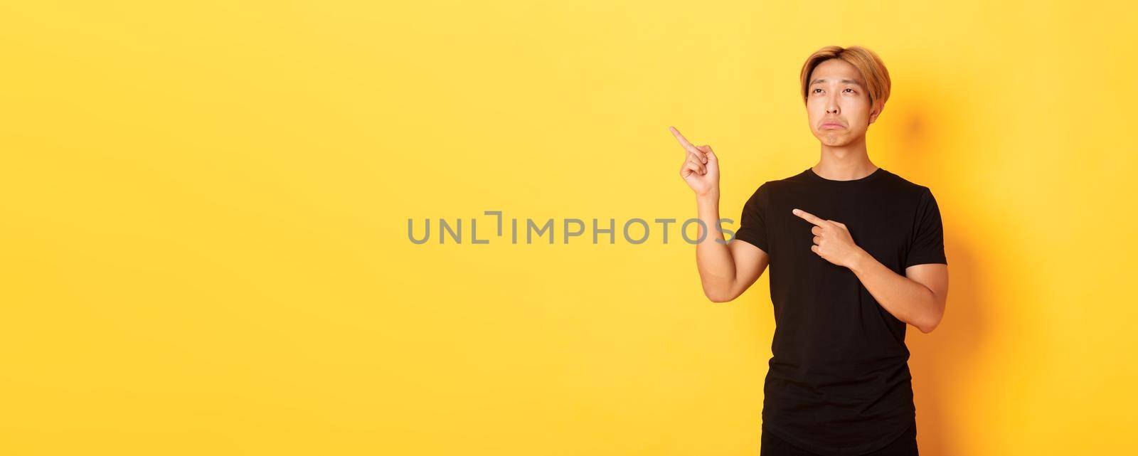 Portrait of pouting upset and funny asian guy pointing and looking upper left corner with regret, standing yellow background by Benzoix