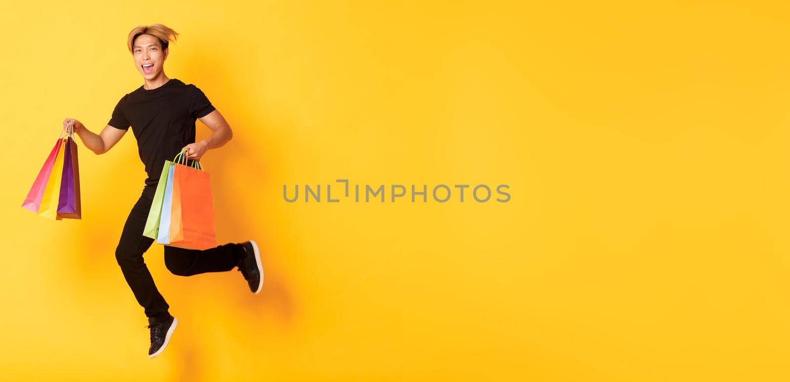 Full-length of happy attractive asian man jumping from happiness and carry shopping bags, standing yellow background by Benzoix