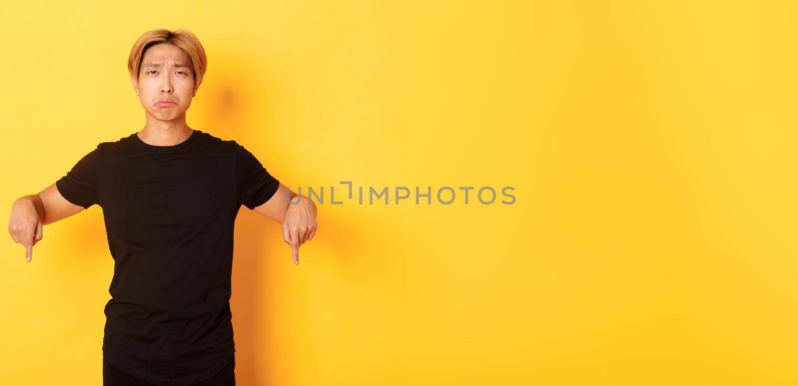 Portrait of disappointed gloomy asian guy, pouting upset and pointing fingers down with regret and uneasy feeling, yellow background.
