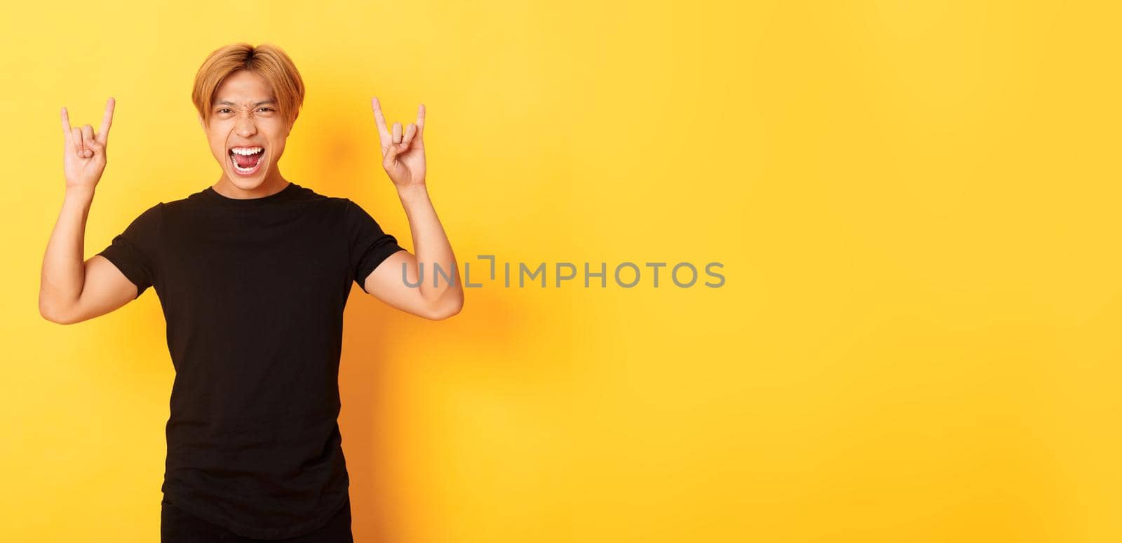 Portrait of happy handsome asian man having fun and partying, showing rock-n-roll gesture, standing yellow background by Benzoix