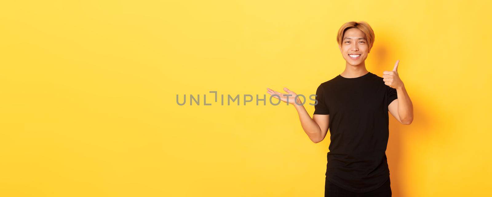 Satisfied and happy attractive korean guy smiling, showing thumbs-up in approval with rejoice, holding something on hand, standing yellow background.