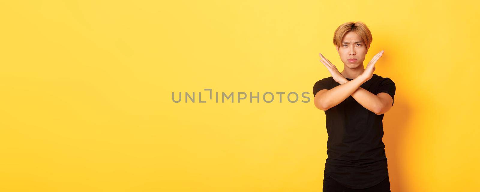 Portrait of disappointed frowning asian man, making cross gesture to stop something bad, standing yellow background.