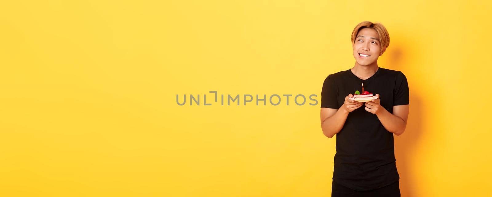 Portrait of handsome dreamy asian guy looking upper left corner and thinking, making wish while celebrating birthday and holding b-day cake, yellow background by Benzoix