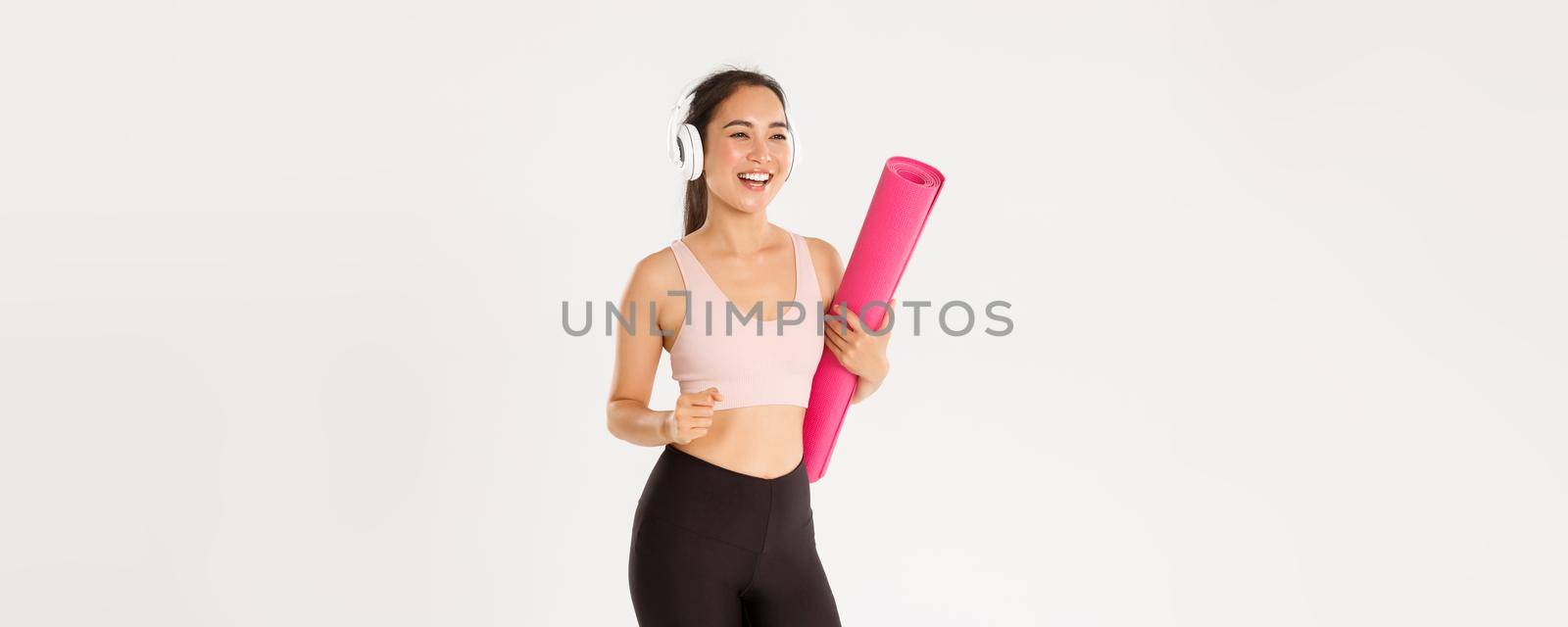 Sport, wellbeing and active lifestyle concept. Cheerful smiling asian fitness girl in headphones and sportswear going to gym with rubbermat, hurry up for yoga classes excited, white background.