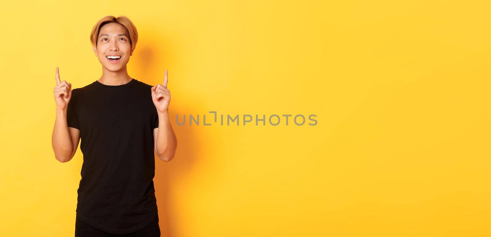 Portrait of satisfied asian blond guy looking amazed, smiling and pointing fingers up, standing yellow background by Benzoix