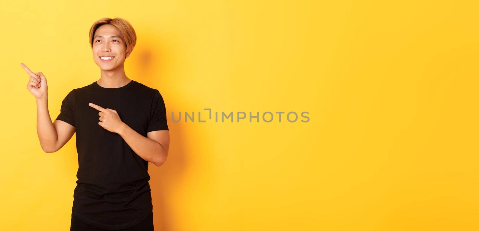 Portrait of handsome korean blond guy, pointing fingers upper left corner and looking satisfied with pleased smile, yellow background by Benzoix