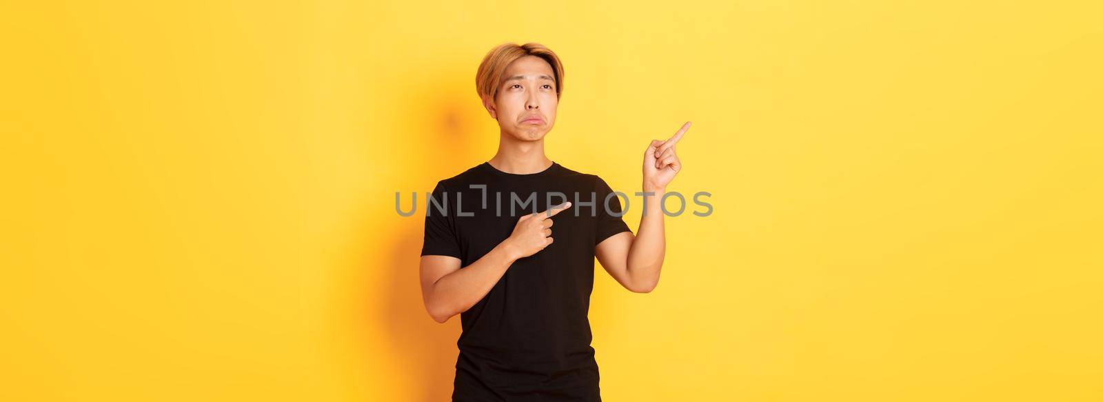 Portrait of pouting upset and funny asian guy pointing and looking upper left corner with regret, standing yellow background.
