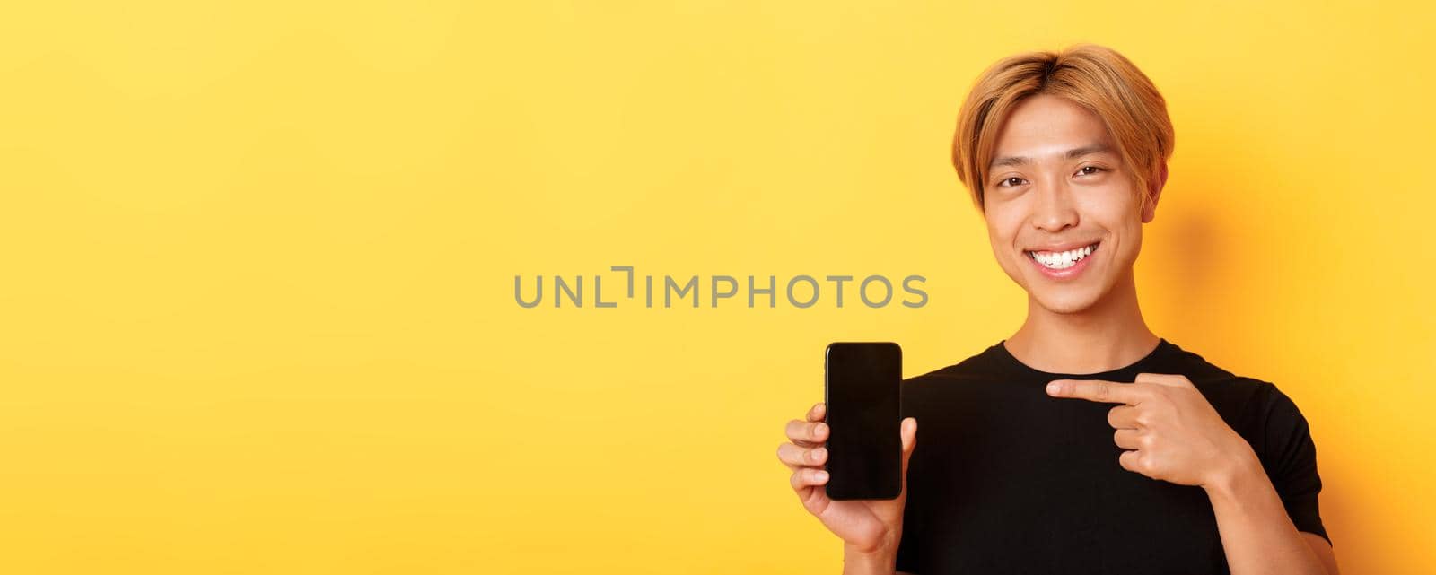 Cheerful attractive korean guy pointing finger at mobile phone screen, smiling pleased, standing over yellow background.
