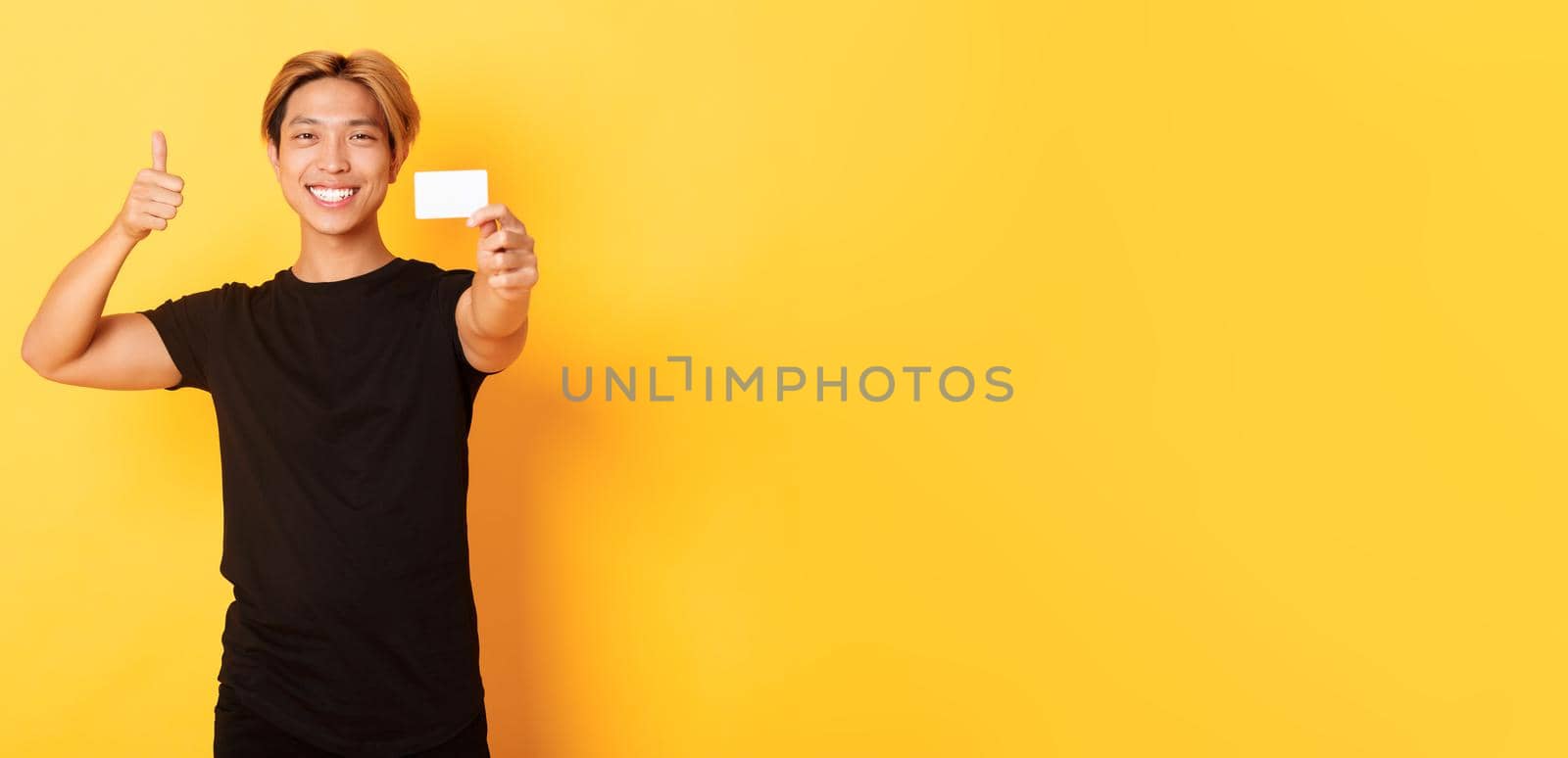 Satisfied handsome asian guy smiling pleased and showing credit card, thumbs-up in approval, standing yellow background by Benzoix