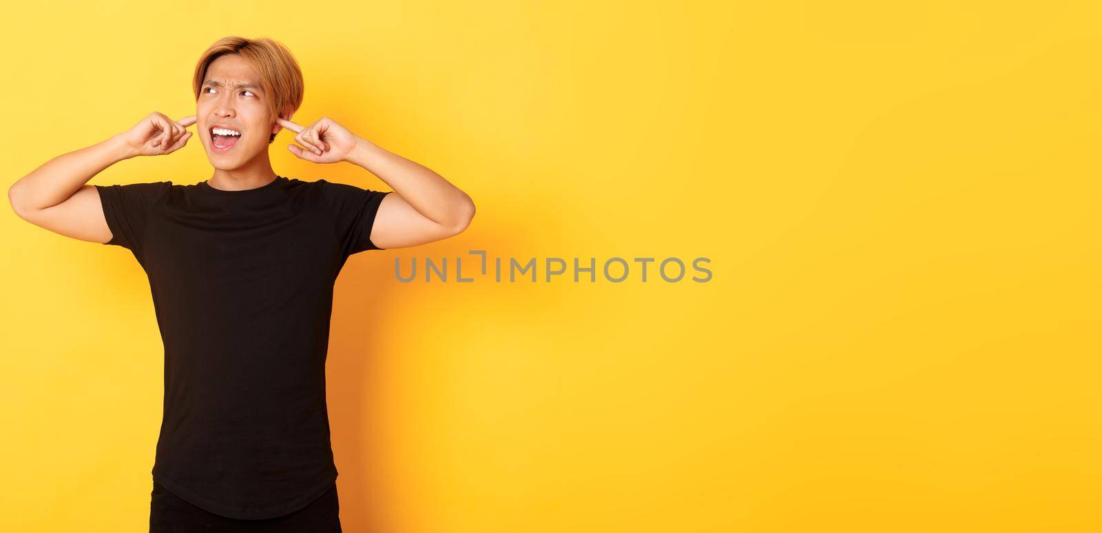 Portrait of annoyed and bothered asian guy grimacing, shut ears disturbed by loud noise, standing yellow background.